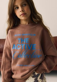 Hnedá mikina s modrým grafickým textom "RESORT SPORTS CLUB THE ACTIVE Collective." Má voľný strih a rebrovaný výstrih.