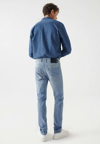 Svijetloplave traperice slim fit kroja, s pet džepova i crnom kožnom oznakom na stražnjem struku. Nosene su sa bijelim tenisicama.