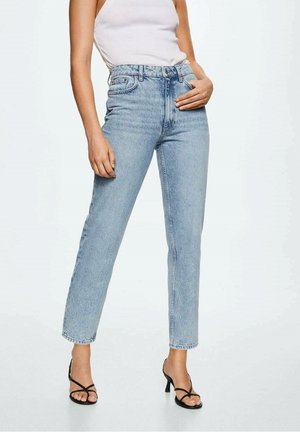 Osoba ubrana w jasnoniebieskie dżinsy z wysokim stanem, biały bezrękawnik i czarne sandały na obcasie z paskami, stojąca z jedną ręką na talii.