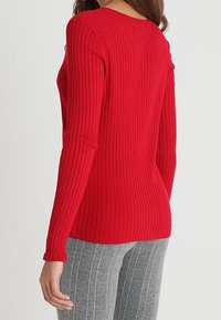 Donna che indossa un top rosso aderente a maniche lunghe a coste con pantaloni grigi a righe, in piedi con la schiena rivolta verso la macchina fotografica.