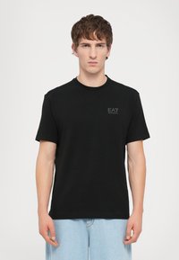 Juoda trumpomis rankovėmis medvilninė marškinėliai suapvalintu kaklu ir subtiliu EA7 logotipu viršutiniame kairiajame kampe.
