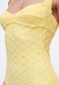 Κοντινό πλάνο ενός κίτρινου αμάνικου φορέματος με υφή κεντήματος με ανθικά μοτίβα και εφαρμοστή κορυφή σε ένα άτομο.