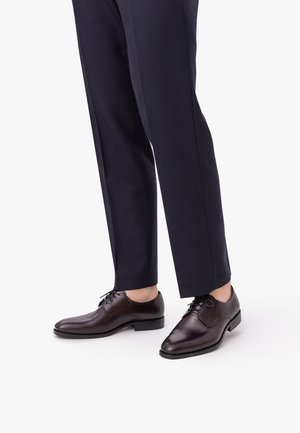 Meeste tumepruunid nahast kleitkingad kantud mereväe siniste mõõtmispükstega, esitatud valgel taustal.