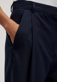 Dunkelblaue Hosen aus einem weichen, strukturierten Stoff zeichnen sich durch ein ordentlich gefaltetes Design mit einer Seitentasche aus, was den maßgeschneiderten Look unterstreicht.
