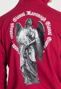 Červená mikina s veľkým čierno-bielym grafickým zobrazením anjela na chrbte, okolo ktorého je text "Gianni Kavanagh" v štýlovom písmovom prevedení.