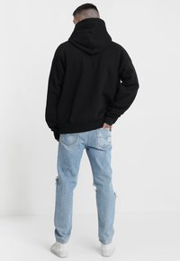 Svart huva sweatshirt med en lös passform, kombinerad med ljusblå slitna jeans och vita sneakers, sedd bakifrån.