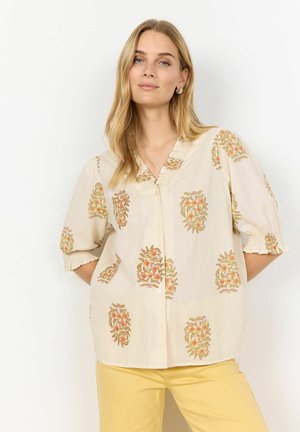 Blouse à manches courtes avec boutons, fabriquée en tissu léger. Fond crème avec des motifs floraux en orange et vert. Manches à poignets froncés.