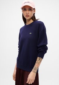Námorníckomodrý sveter s okrúhlym výstrihom vyrobený z pleteniny, s malým zeleným logom na ľavej hrudi a dlhými rukávmi. Model má na sebe ružovú čiapku.