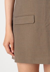 Hnědá sukně s kostkovaným vzorem houndstooth, s rovnými liniemi, která má jednu lištovou kapsu a hladkou texturu.
