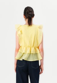 Mujer con el cabello oscuro recogido en un moño, vestida con una blusa amarilla sin mangas con volantes y jeans azul oscuro, de pie contra un fondo blanco.