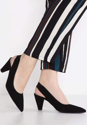 Stopy w czarnych zamszowych sandałkach slingback na słupku, zestawione z czarnymi spodniami z pionowymi białymi, niebieskimi i brązowymi paskami.