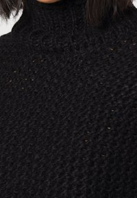 Schwarzer gestrickter Pullover mit hohem Kragen und grober Textur sowie einem offenen Maschenmuster. Der Stoff wirkt dick und gemütlich.