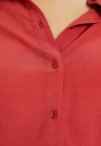 Camisa roja de botonadura frontal hecha de una tela suave, que presenta un cuello, dos botones y un único bolsillo frontal. Textura suave con un ligero brillo.