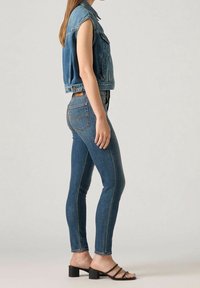 Chaleco de mezclilla sin mangas con un ajuste ligeramente recortado, combinado con jeans ajustados de color azul oscuro. Sandalias negras de tacón con tirantes completan el look.