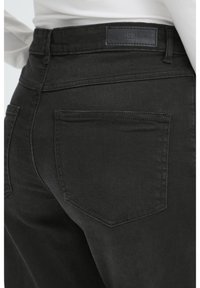 Detailný záber na zadné vrecko a pás čiernych džínsov z denimu, nosených s bielym topom, zobrazujúci šitie a koženú značkovú etiketu.