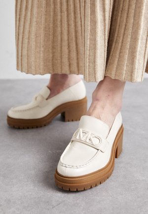 Mocasines de cuero crema con tacones marrones gruesos y suelas de goma, combinados con pantalones beige de punto acanalado.