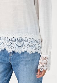 Witte lange mouw blouse met ingewikkelde kantafwerking aan de zoom en manchetten, met een zachte textuur en een ontspannen pasvorm, gedragen met blauwe jeans.