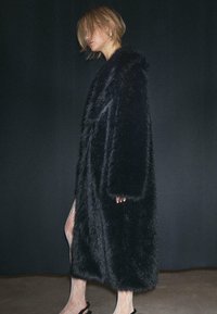 Crni kaput od umjetnog krzna s širokim kragnom, dugim rukavima i širokim krojem, prikazan protiv tamne pozadine. Silueta se kontrastira s prozirnim tajicama.