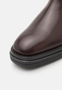 Zapato de cuero marrón oscuro, suave, con punta redonda y suela negra en capas sobre fondo blanco.