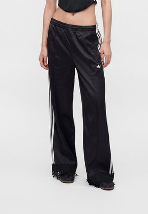 Γυναίκα που φοράει μαύρο σατέν παντελόνι Adidas με λευκές ρίγες και δαντέλα στο τελείωμα, σε συνδυασμό με μαύρα παπούτσια και cropped μαύρο τοπ.