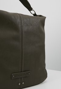Sac à main en cuir vert olive avec une texture lisse, une couture argentée et une plaque de marque. Dispose d'une seule poignée pour le transport.