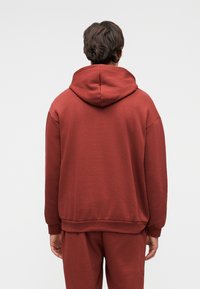Homme debout de dos portant un sweat à capuche couleur rouille et un pantalon de survêtement assorti, sur un fond clair uni.