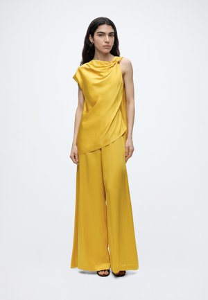 Donna con lunghi capelli scuri che indossa un top giallo di raso senza maniche e pantaloni a gamba larga, in piedi su uno sfondo bianco semplice.