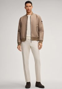 Giacca bomber color beige con finitura liscia, indossata sopra un maglione bianco. Abbinata a pantaloni beige chiaro e stivaletti alla caviglia in camoscio marrone scuro.