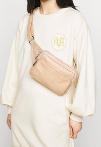 Beige nahkahihnatasku, jossa on kolme vetoketjullista osastoa. Pehmeä pinta ja minimalistinen muotoilu. Kuljetetaan rinnan päällä valkoisen hupparin yli.