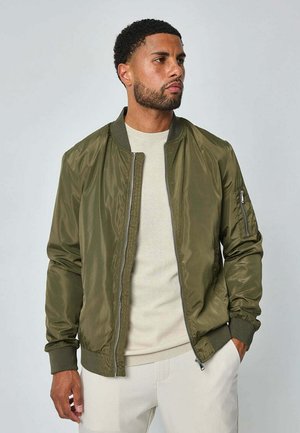 Homme portant une veste bomber vert olive par-dessus un pull beige avec un pantalon clair, debout avec une main dans la poche, regardant vers la droite.