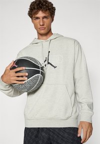 Grå sweatshirt med en lomme foran og snoretræk i hætten, prydet med et sort grafisk logo. Holder en tekstureret sort basketball med grå accenter.