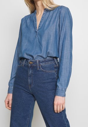 Vrouw die een blauw, lang mouw, V-halsblouse draagt, ingestopt in een high-waist donkere blauwe jeans, staand tegen een effen achtergrond.