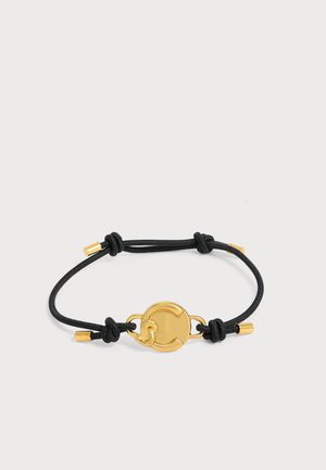 Pulsera de cordón negro con nudos ajustables y un charm circular dorado en el centro, con remates metálicos dorados en cada extremo, sobre fondo blanco.