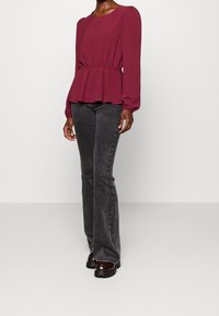 Kvinna iförd en långärmad vinbrun blus, svarta högmidjade utsvängda jeans och svarta klumpiga skor framför en enkel bakgrund.