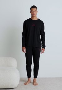 Cămașă neagră cu mânecă lungă, detaliu roșu al logo-ului, asociată cu pantaloni negri tapered. Țesătura pare moale, potrivită pentru haine de relaxare.