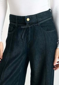 Denim broek in donkerblauw met zichtbare stiksels, een taille met riem, zijzakken en een losse pasvorm. Gladde textuur met een lichte glans.