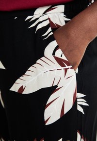 Černá látka s velkými bílými listy a hnědými akcenty. Detailní záběr na ruku ve boční kapse, který zdůrazňuje design oděvu.