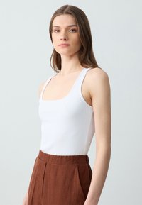 Weißes geripptes Tanktop mit quadratischem Ausschnitt, kombiniert mit braunen, hochgeschnittenen Leinenhosen mit gefalteter Vorderseite und elastischem Bund.
