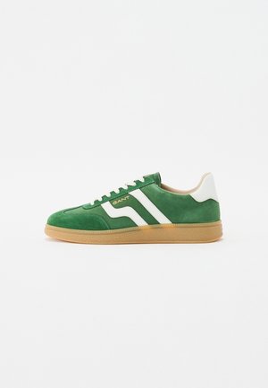 Grønn semsket sneaker med hvite striper og snører, med en gummirådgummisåle. Merking på siden i gullskrift. Klassisk design.