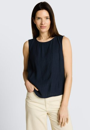 Frau mit langen braunen Haaren, die ein ärmelloses dunkelblaues Top und hellbeige Hosen trägt, eine Hand in der Tasche, neutraler Hintergrund.