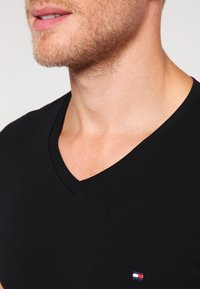 Czarna bawełniana koszulka V-neck z małym logo w czerwono-białoniebieskich kolorach na dolnym lewym boku; gładka tekstura i dopasowany krój.