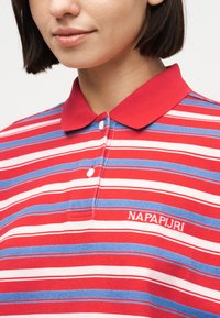 Κοντινή λήψη ενός ατόμου που φοράει πόλο μπλούζα με κόκκινες, λευκές και μπλε ρίγες, με κόκκινο γιακά και το κέντημα "NAPAPIJRI" στο στήθος.