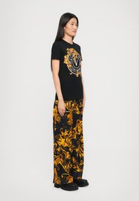 Woman stands sideways wearing black T-shirt with gold ornate logo and matching wide-leg pants with gold baroque pattern, black chunky shoes.