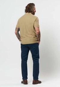 Homme aux cheveux bouclés et tatouages, tourné de dos, portant un polo beige, un pantalon bleu marine et des chaussures marron sur un fond uni.