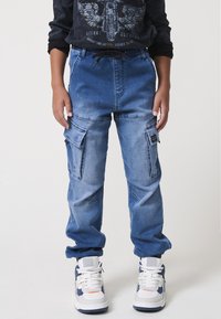 Gradient kék farmer cargo nadrág, több oldalsó zsebbe, rugalmas derékkal és mandzsettával; fehér és tengerészkék sportcipőkkel párosítva.