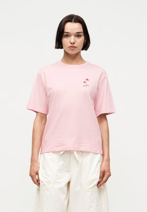 Γυναίκα με κοντά σκούρα μαλλιά, που φοράει ροζ ανοιχτό T-shirt με μικρά κόκκινα λουλουδάκια και φαρδιά άσπρα παντελόνια, στέκεται μπροστά σε απλό φόντο.