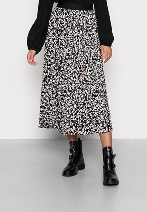 Vrouw draagt een geplooide midi-rok met een abstracte print in zwart, wit, beige en blauw, gecombineerd met zwarte leren enkellaarsjes met gespen.
