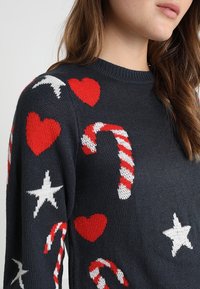 Stickad marinblå tröja med röda hjärtan, vita stjärnor och godisstångsmönster. Klassisk rundhalsdesign med en texturerad finish.