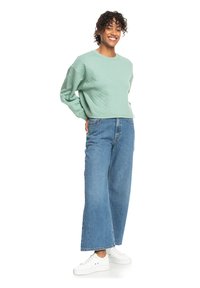 Felpa trapuntata verde menta con una vestibilità rilassata, abbinata a jeans blu a vita alta e gamba ampia, e scarpe da ginnastica bianche.