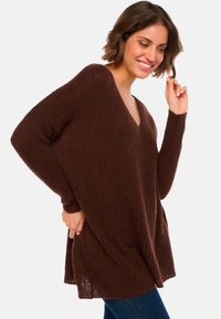 Pull en tricot brun, ample, avec un col V, texture côtelée et manches longues. L'ourlet est arrondi sur les côtés, offrant une silhouette décontractée.